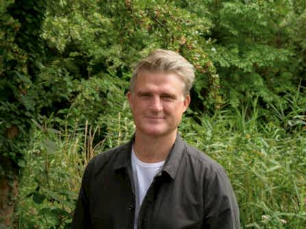 A man standing in front of a green backdrop full of vegetation. The person pictured is David Mooney